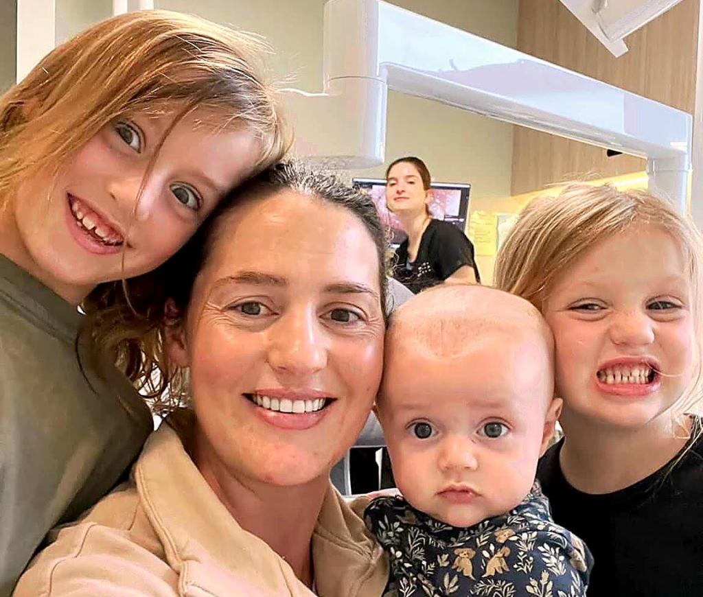 Smiling mom holds a baby while two kids hug the group in an indoor setting, taking a selfie together.